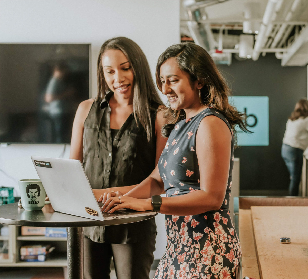 Two women standing at a tall table, working together at a laptop.